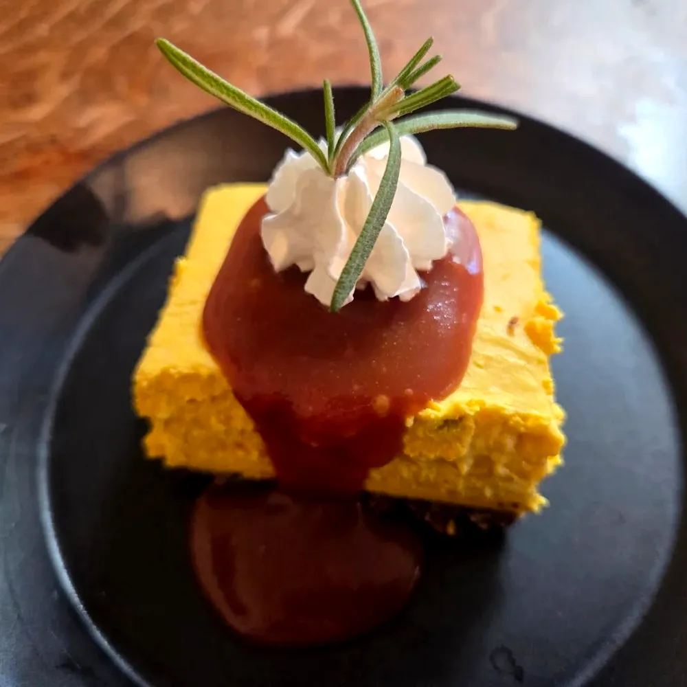 黒い皿にのった鮮やかな黄色のチーズケーキ。ソースとホイップ、ローズマリーが飾られている
